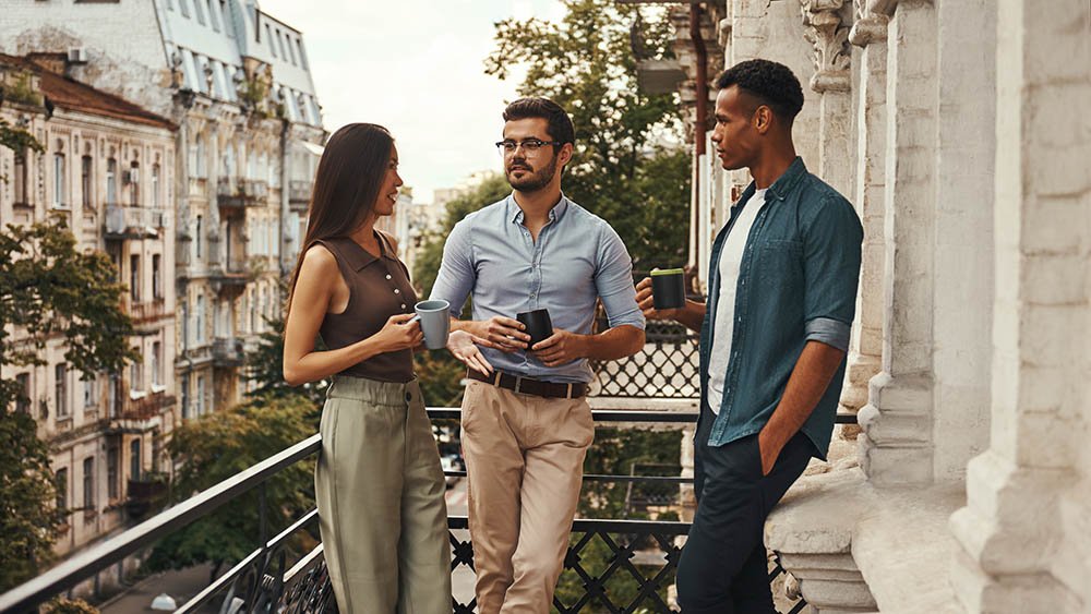 Conversation. Young cheerful colleagues in casual wear holding cups and talking with each other while standing on the balcony. Coworkers resting. Coffee break - properly maintained balcony - by drblcony - balcony inspection